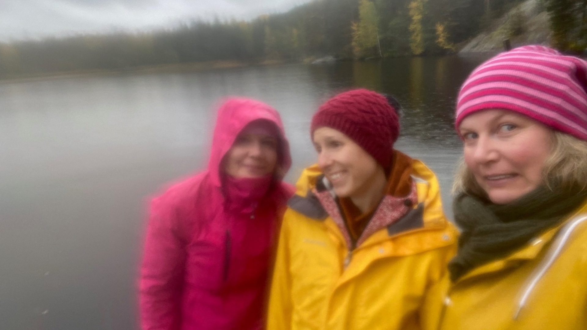 Karikan kolme työntekijää poseraa kameralle sateisen järven edessä sadetakit päällä.